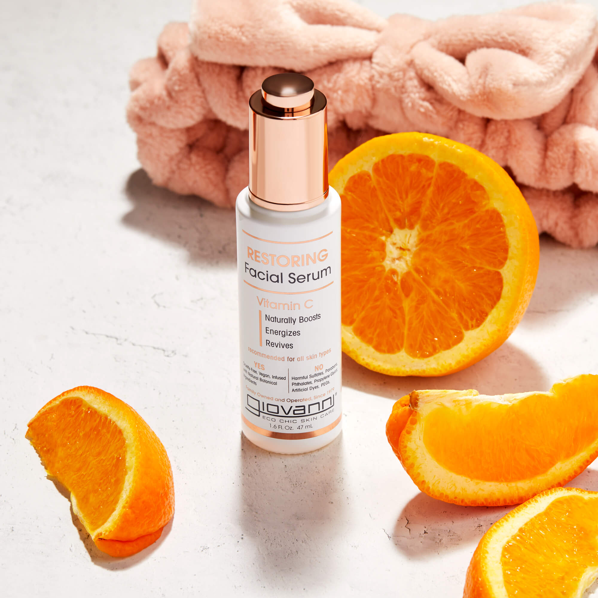A bottle of Giovanni Restoring Facial Serum with Vitamin C stands on a marble surface, surrounded by orange slices and a soft pink headband in the background.