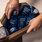 Someone places several blue bottles of Giovanni Cosmetics MEN 2-in-1 Daily Shampoo & Conditioner with Ginseng & Eucalyptus into a brown toiletry bag on a light surface.