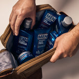 Someone places several blue bottles of Giovanni Cosmetics MEN 2-in-1 Daily Shampoo & Conditioner with Ginseng & Eucalyptus into a brown toiletry bag on a light surface.