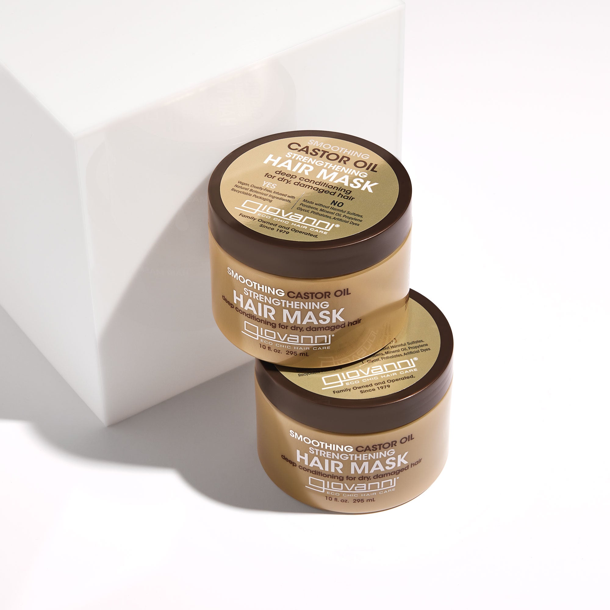 Two jars of Giovanni Cosmetics SMOOTHING CASTOR OIL STRENGTHENING HAIR MASK, in gold packaging with black lids and white text, sit stacked on a white surface—luxuriously hydrating and strengthening dry, damaged hair.