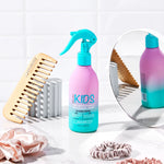 A bottle from the Giovanni Cosmetics KIDs Bundle stands on a counter alongside a wooden wide-tooth comb, scrunchies, and a round mirror, all set against a white tile background.