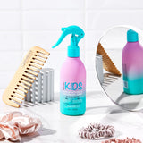 A bottle from the Giovanni Cosmetics KIDs Bundle stands on a counter alongside a wooden wide-tooth comb, scrunchies, and a round mirror, all set against a white tile background.