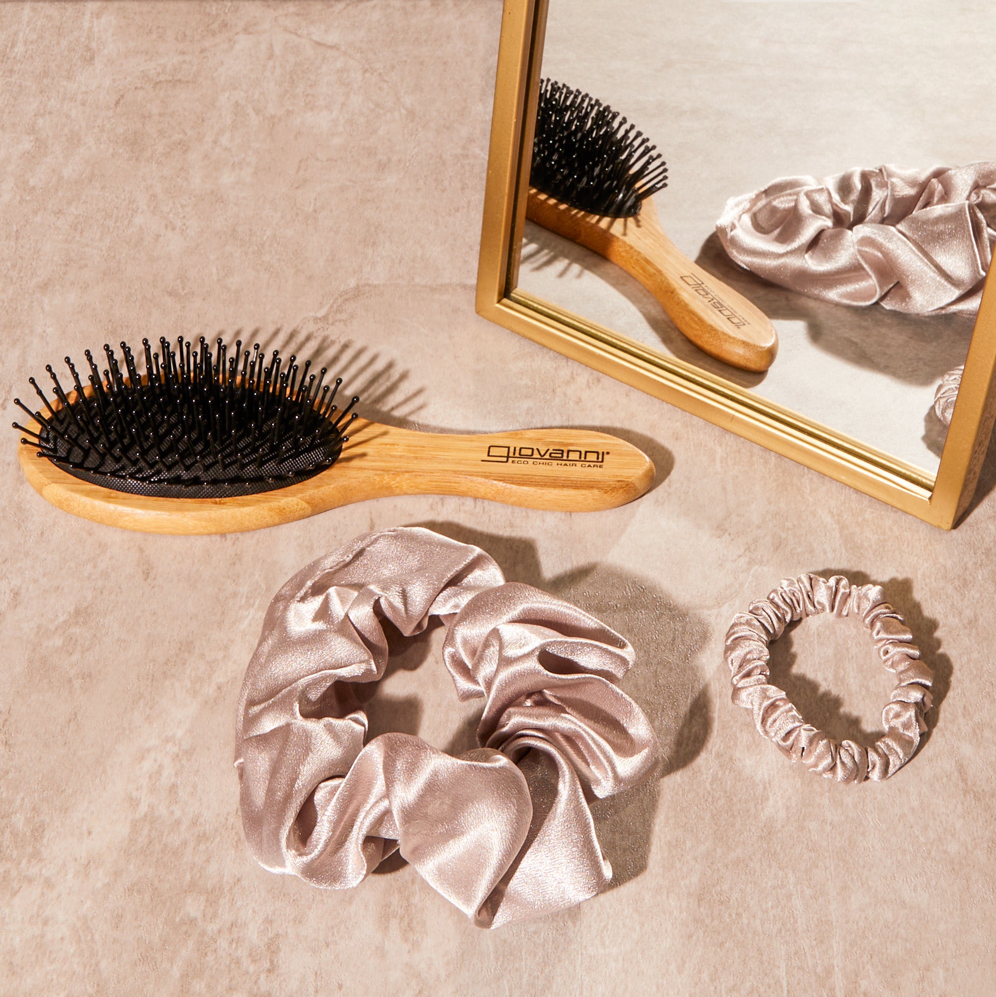 Giovanni Cosmetics' SATIN HAIR SCRUNCHIES (3 pack: Blush, Beige, Dark Brown), a wooden hairbrush, and a mirrored reflection are arranged on a beige surface—ideal for salon-quality hair care at home.