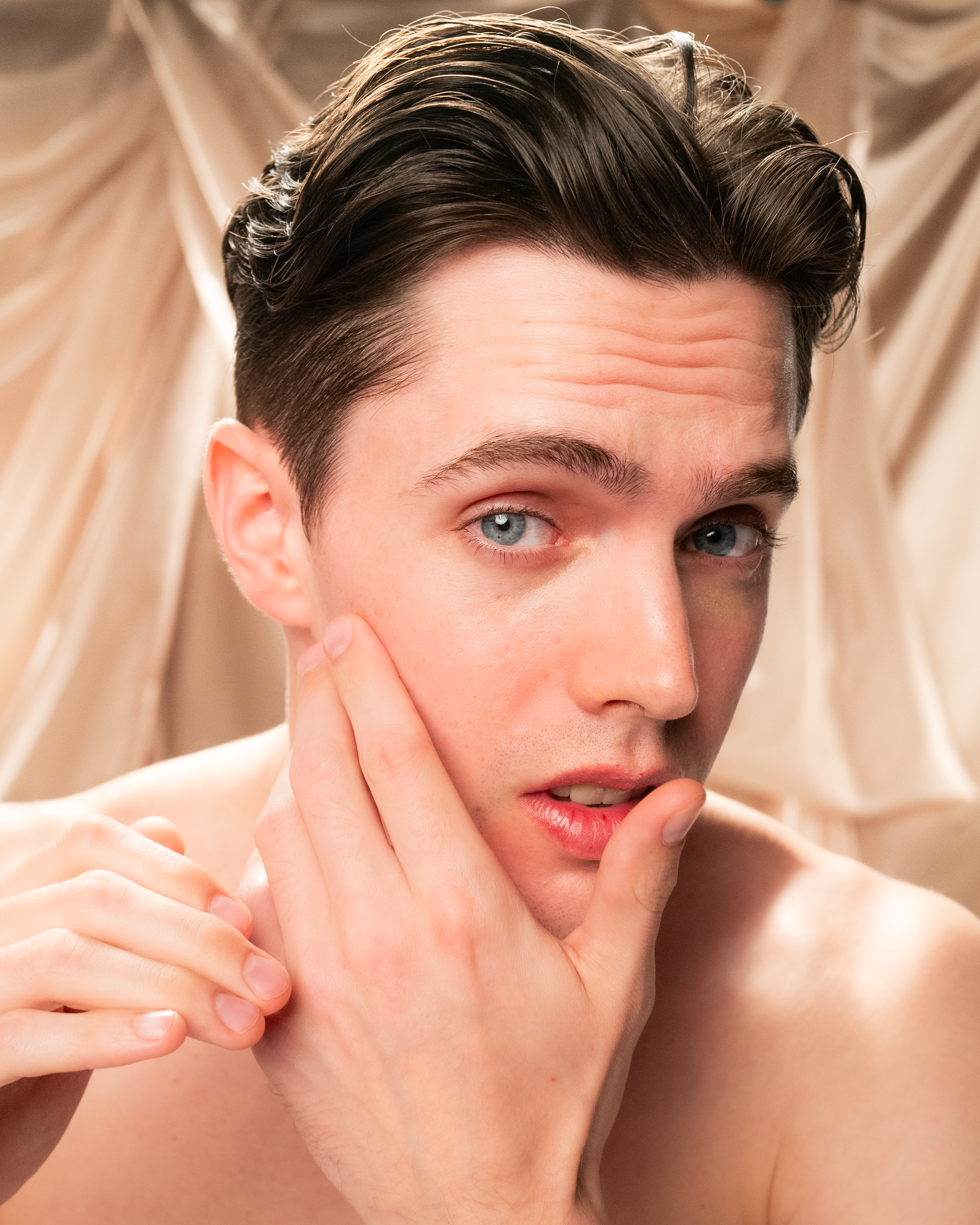 A young man with fair skin and dark hair touches his cheek, looking concerned—perhaps in need of Giovanni Cosmetics' MEN Moisturizing Shave Cream. Soft beige fabric drapes in the background.