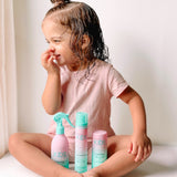 A young child with wet, curly hair smiles in a pink outfit, seated cross-legged. In front are three teal and pink Giovanni Cosmetics ECO CHIC® KIDS hair care products, including the STYLING TAMING & CURL CREAM. The background is bright and softly lit.
