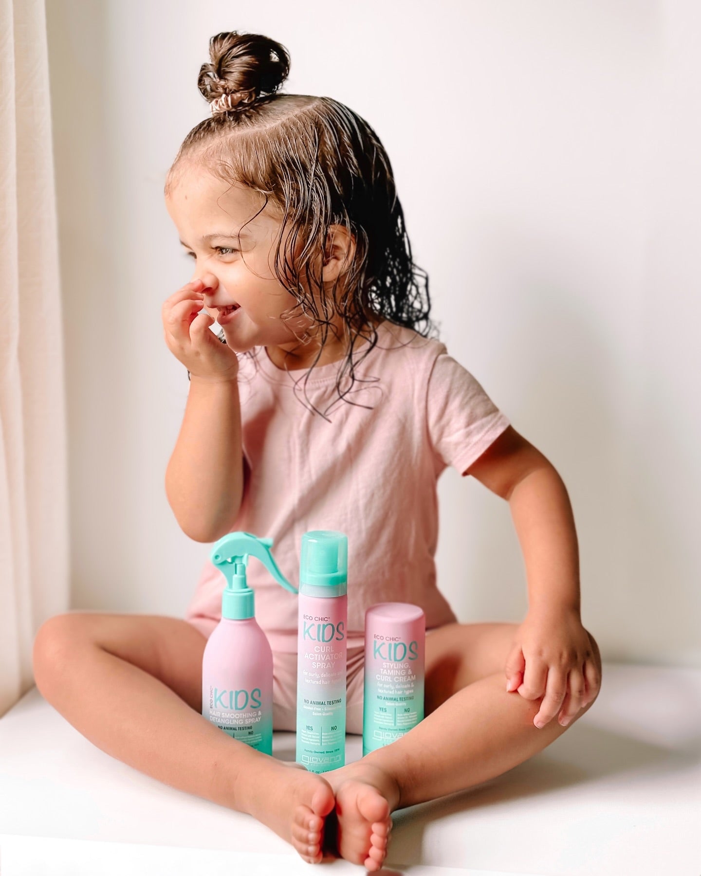 A young child with wet, curly hair smiles in a pink outfit, seated cross-legged. In front are three teal and pink Giovanni Cosmetics ECO CHIC® KIDS hair care products, including the STYLING TAMING & CURL CREAM. The background is bright and softly lit.