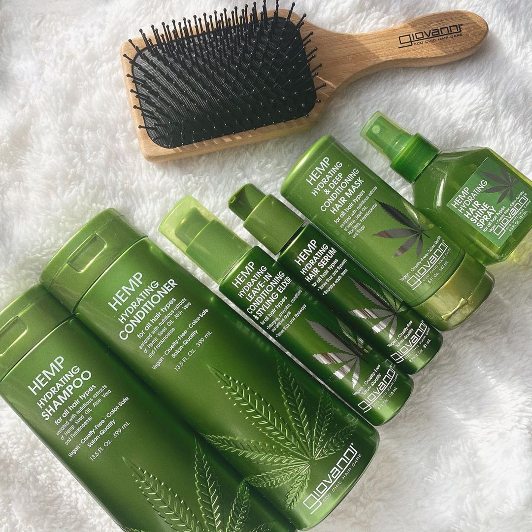 Six green Giovanni Cosmetics HEMP HYDRATING LEAVE-IN CONDITIONING & STYLING ELIXIR bottles, featuring hemp leaf graphics for hydrating, color-safe hair care, are arranged with a wooden brush on a white fluffy surface.