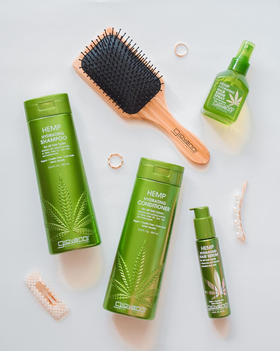 Flat lay of Giovanni Cosmetics HEMP HYDRATING HAIR SERUM in a green bottle, styled with a wooden hairbrush, hair clips, and a ring on a light surface.
