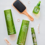 Flat lay of Giovanni Cosmetics HEMP HYDRATING HAIR SERUM in a green bottle, styled with a wooden hairbrush, hair clips, and a ring on a light surface.